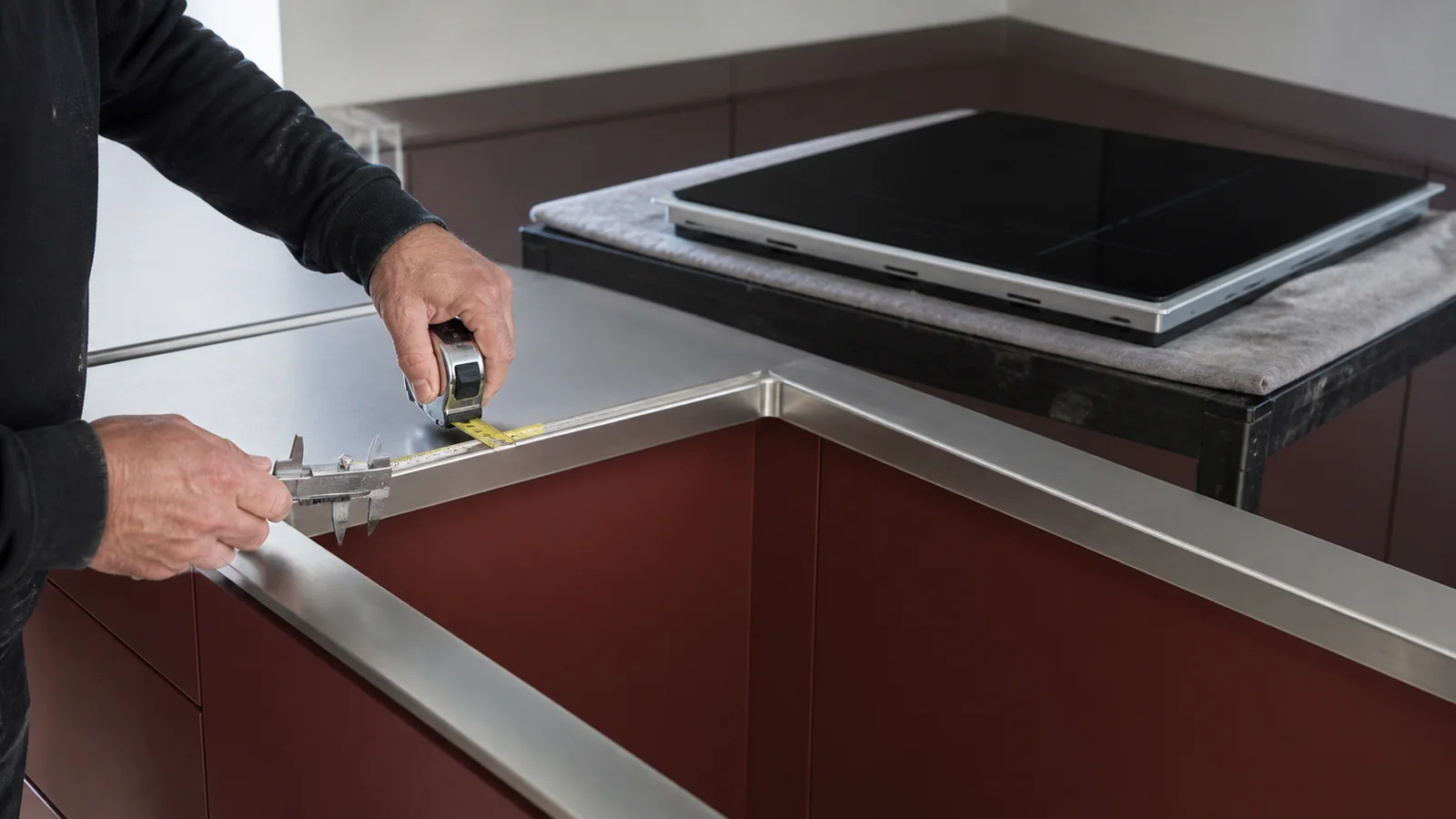 Installer measuring the finished metal return around a cooktop cutout while a replacement induction cooktop waits nearby